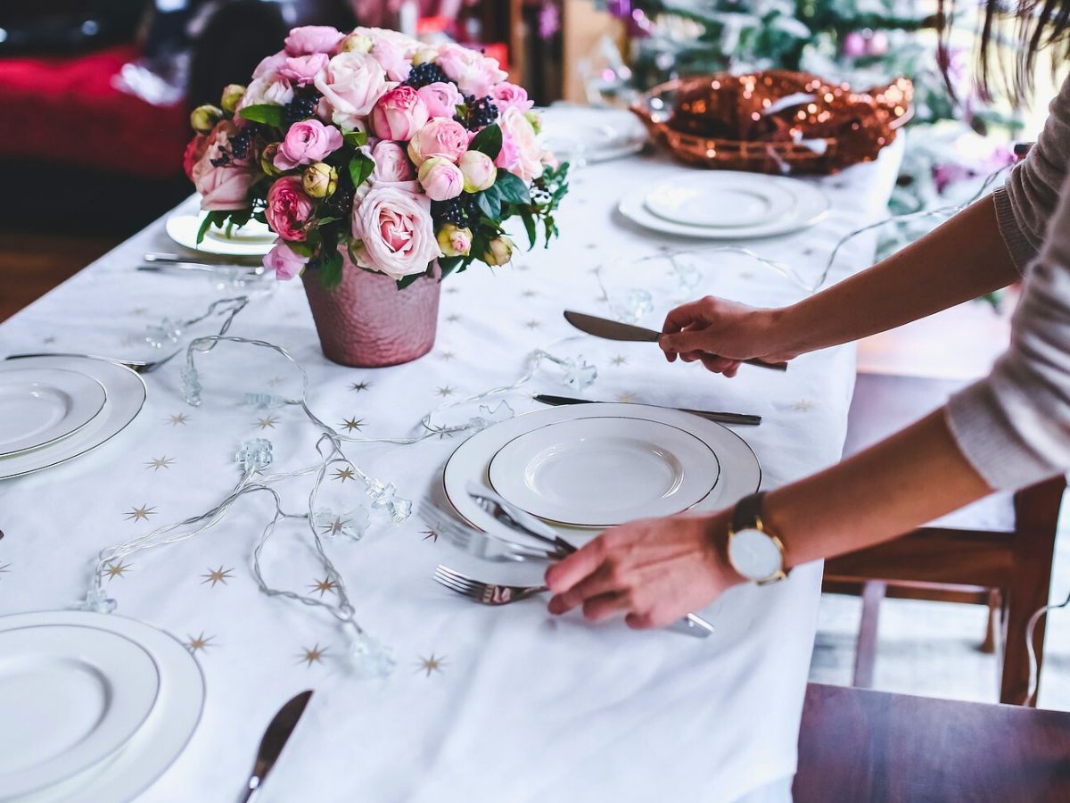 A woman setting a dining table with plates and silverware, adorned with a flower centerpiece and string lights.