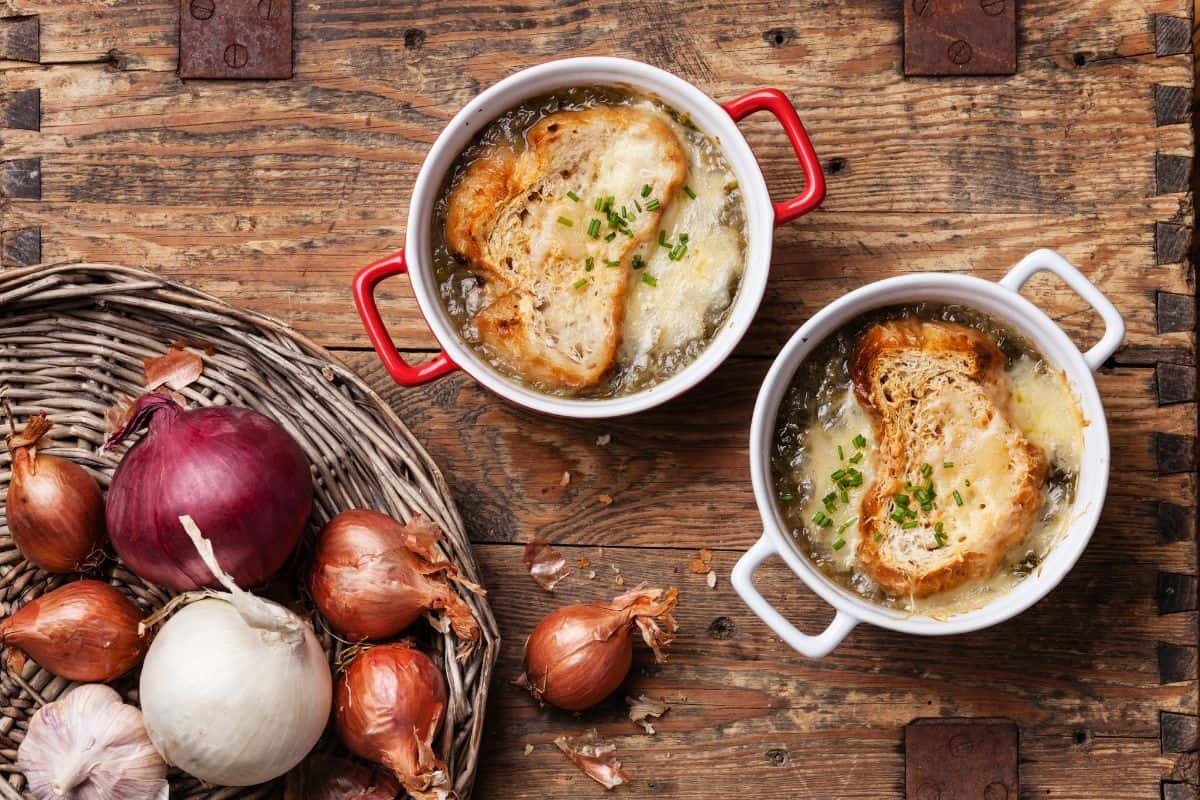 Bowls of French onion soup on wooden table next to a bowl of onions