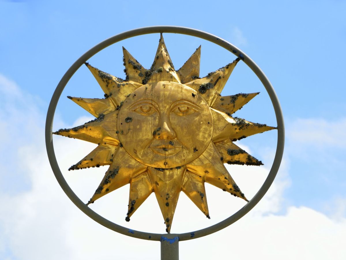 Golden sun sculpture with a face, set against a blue sky, encircled by a metal ring.