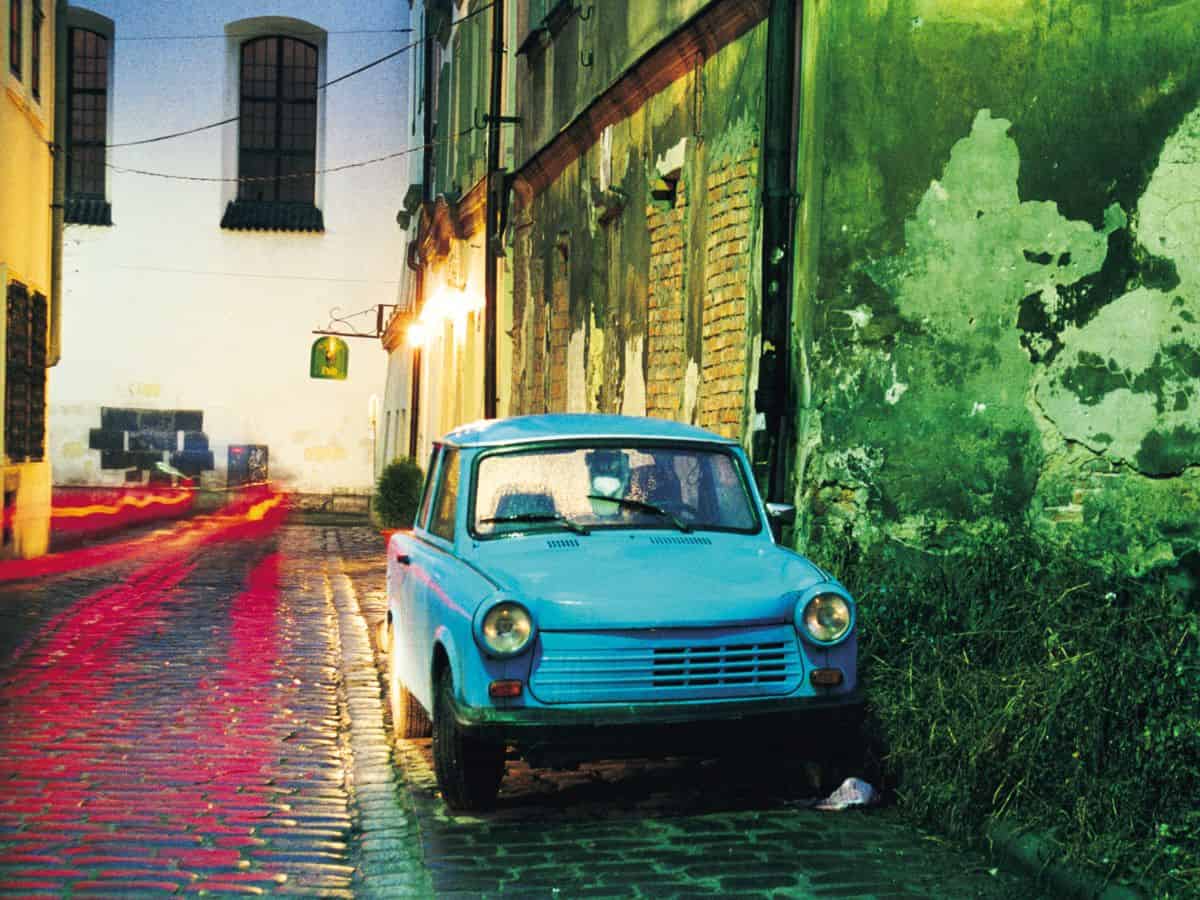 A blue Trabant car parked on a cobblestone street in the rain.