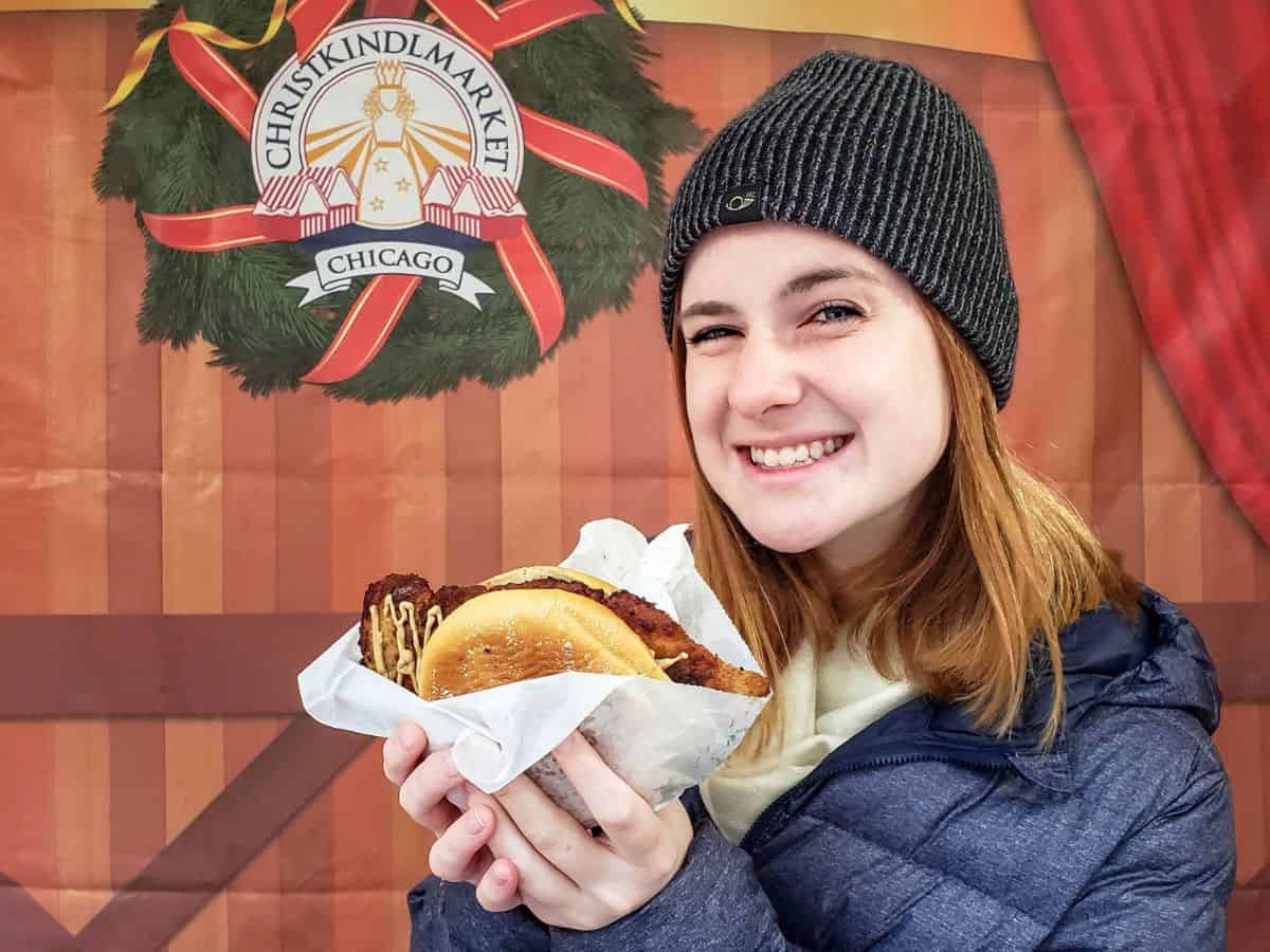 A girl holding a schnitzel sandwich in front of a Christkindlmarket banner in Chicago.