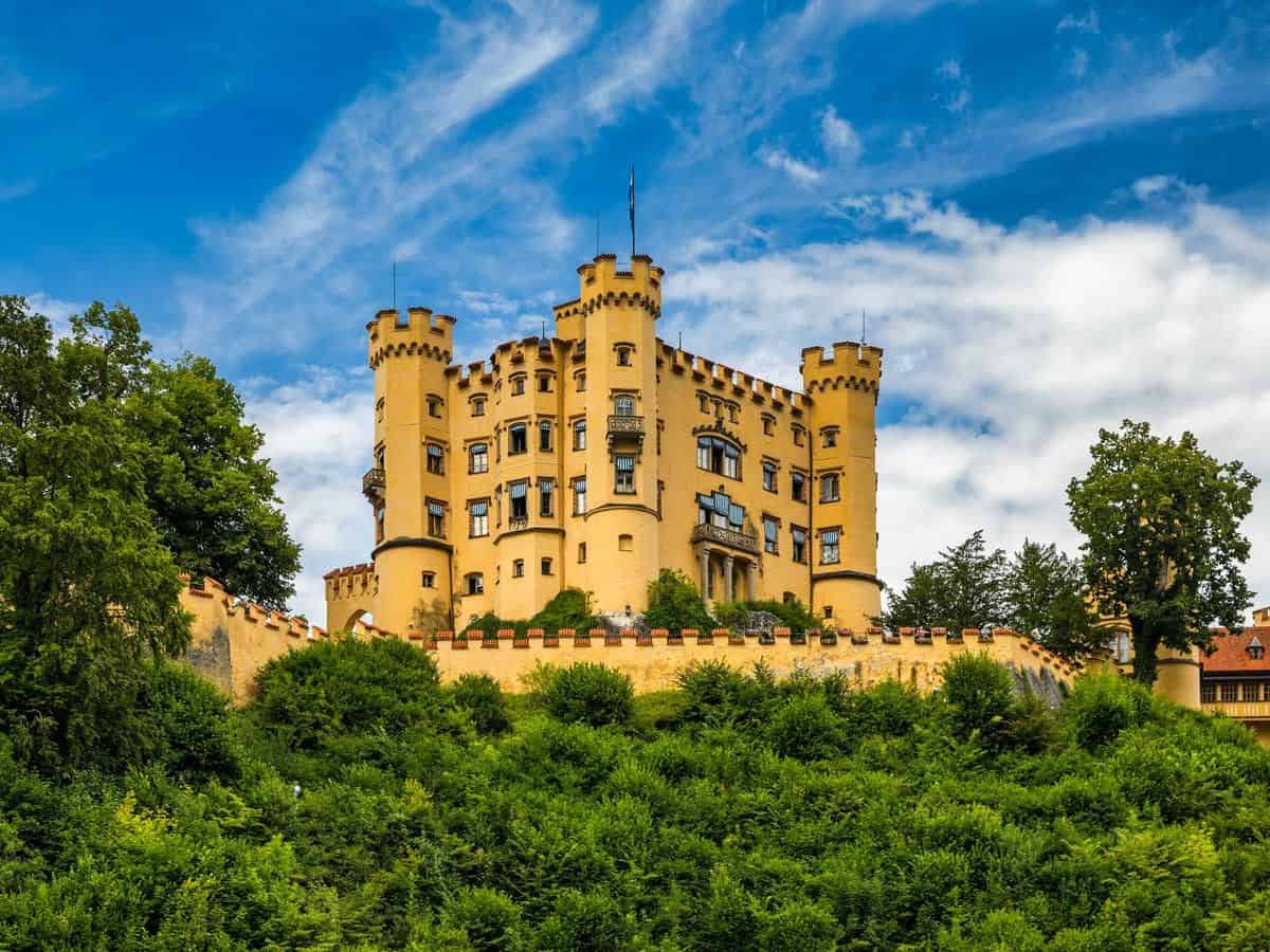 Hohenschwangau Castle sits on top of a green hill in Germany. castles in Germany, sits on top of a green hill.
