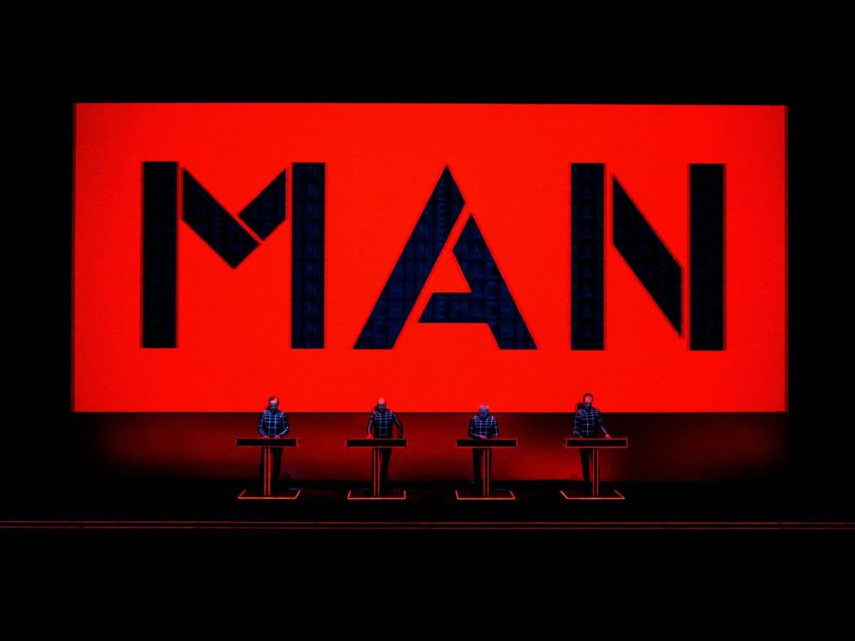 Kraftwerk performing on stage in front of a big, red sign that reads, "Man."