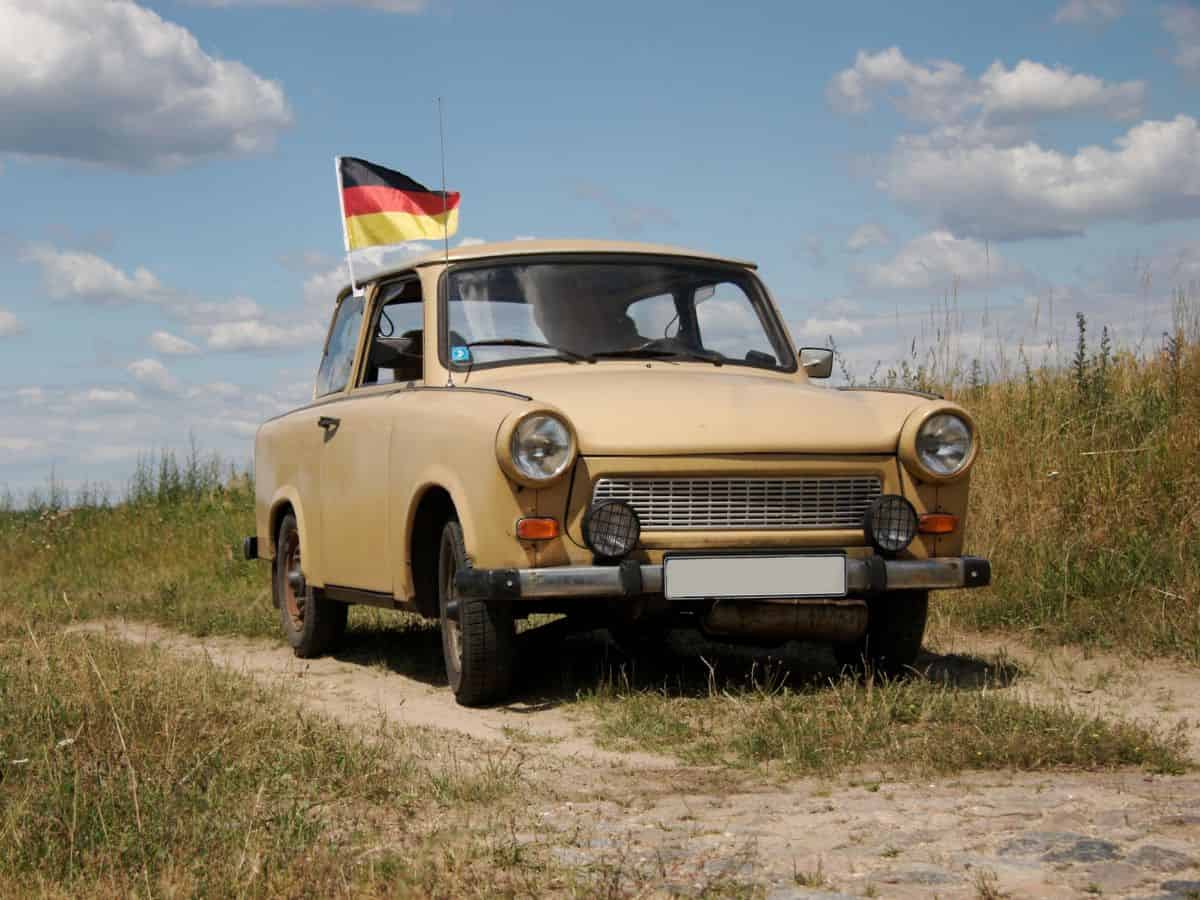 An old Trabant car with a German flag parked on a dirt road.