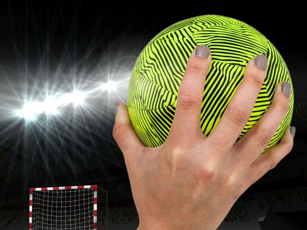 A woman's hand holding a yellow handball in front of a goal.