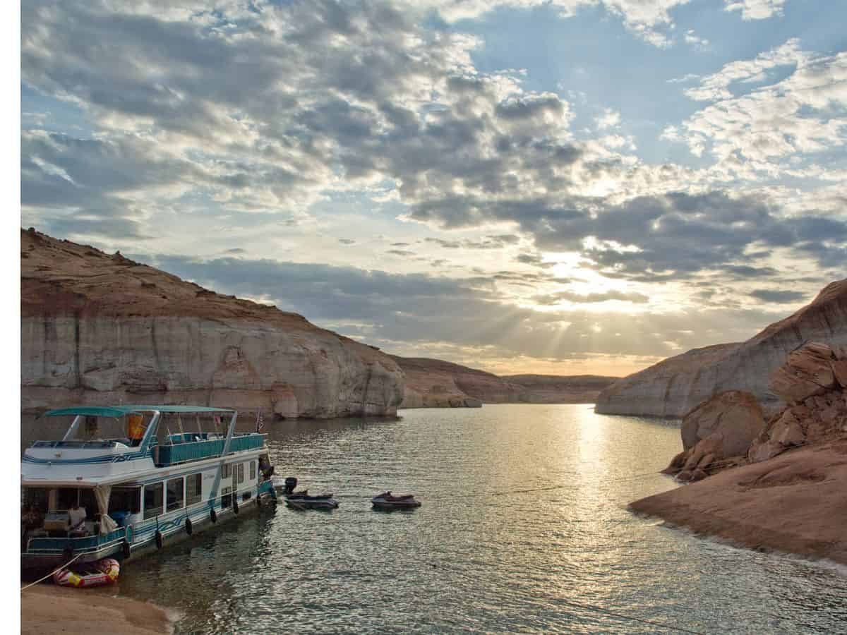 A houseboat docked along the bank of Lake Powell surrounded by people out in the calm, clear water.