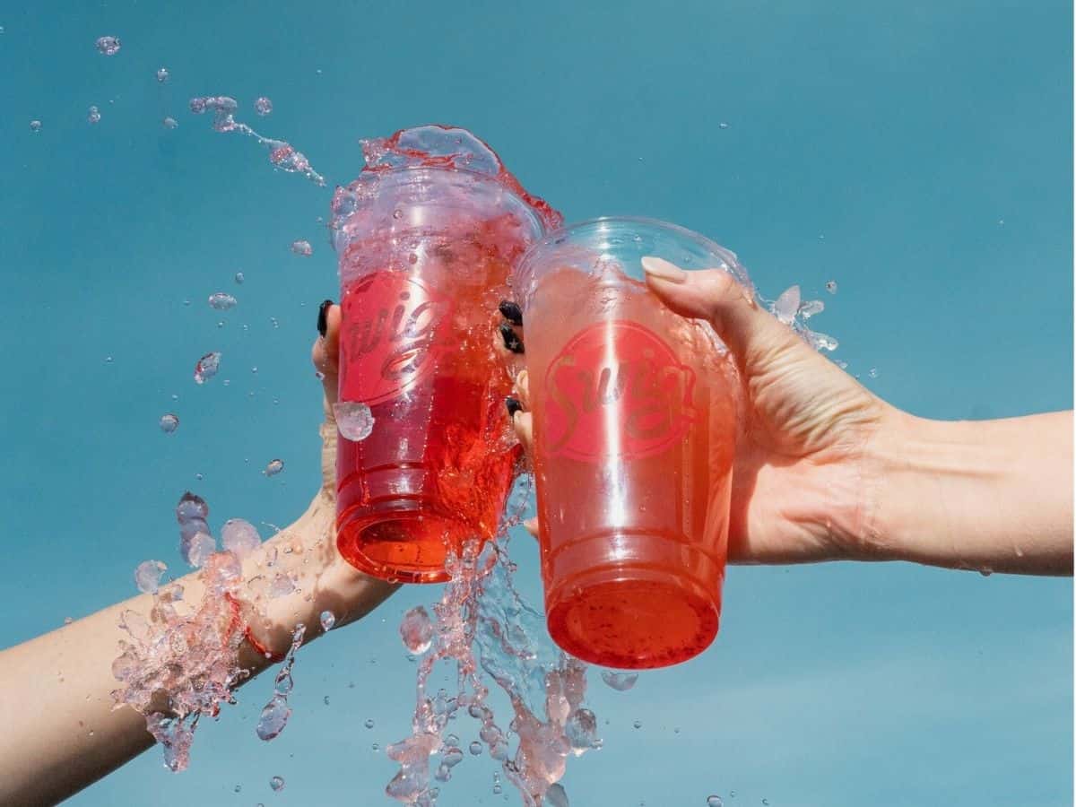 Two hands holding two plastic cups with specialty soda in them.
