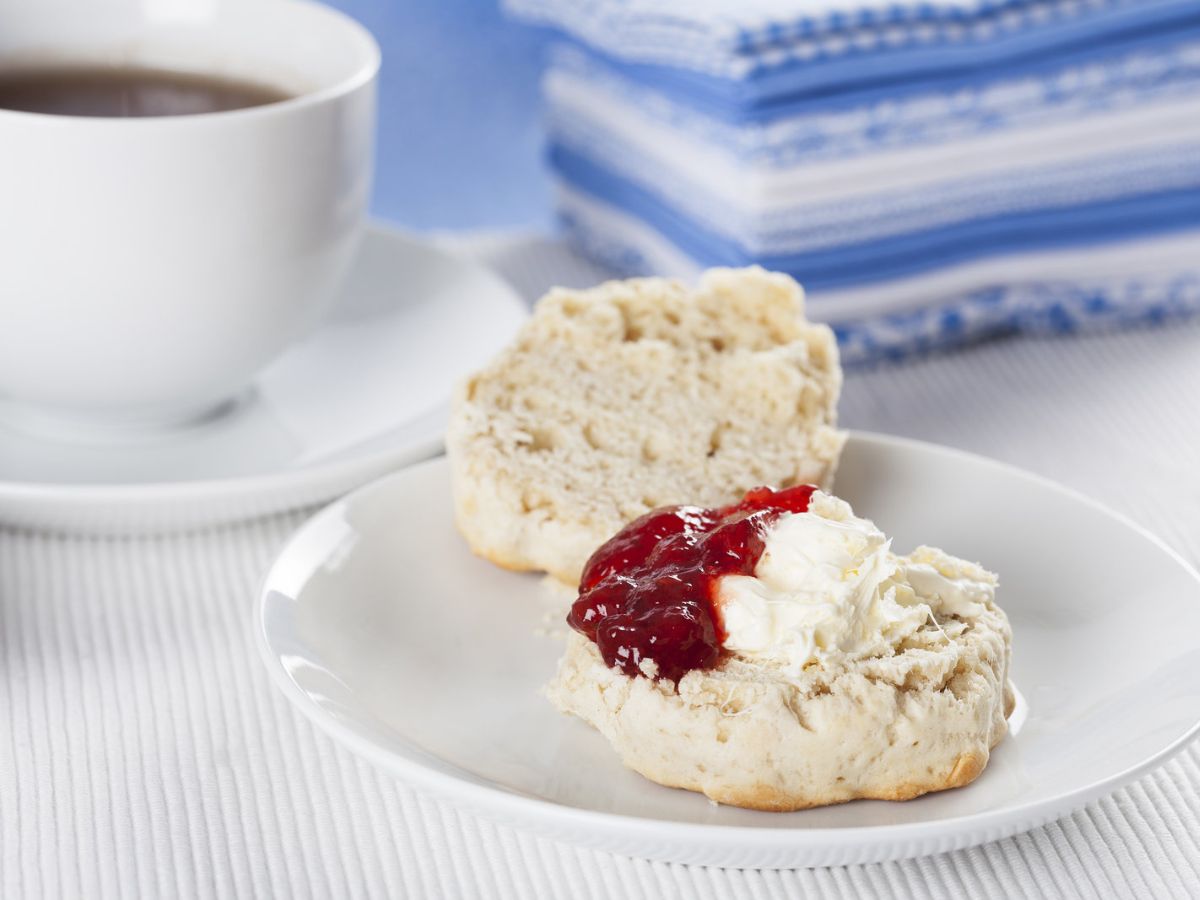 A halved scone topped with cream and jam next to a cup of tea.