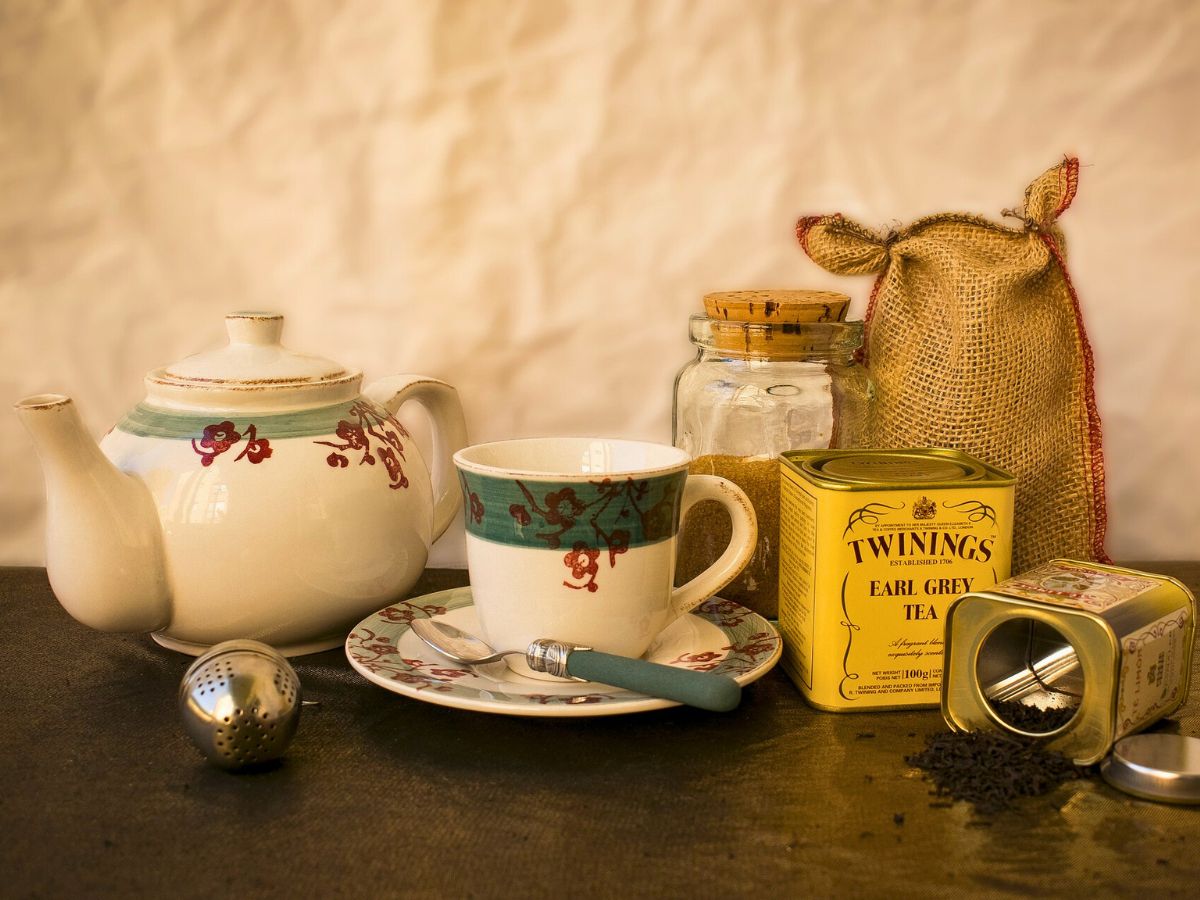 A still life arrangement of tea time essentials, including a porcelain teapot, cup and saucer, a tin of earl grey tea, a strainer, and loose tea leaves.