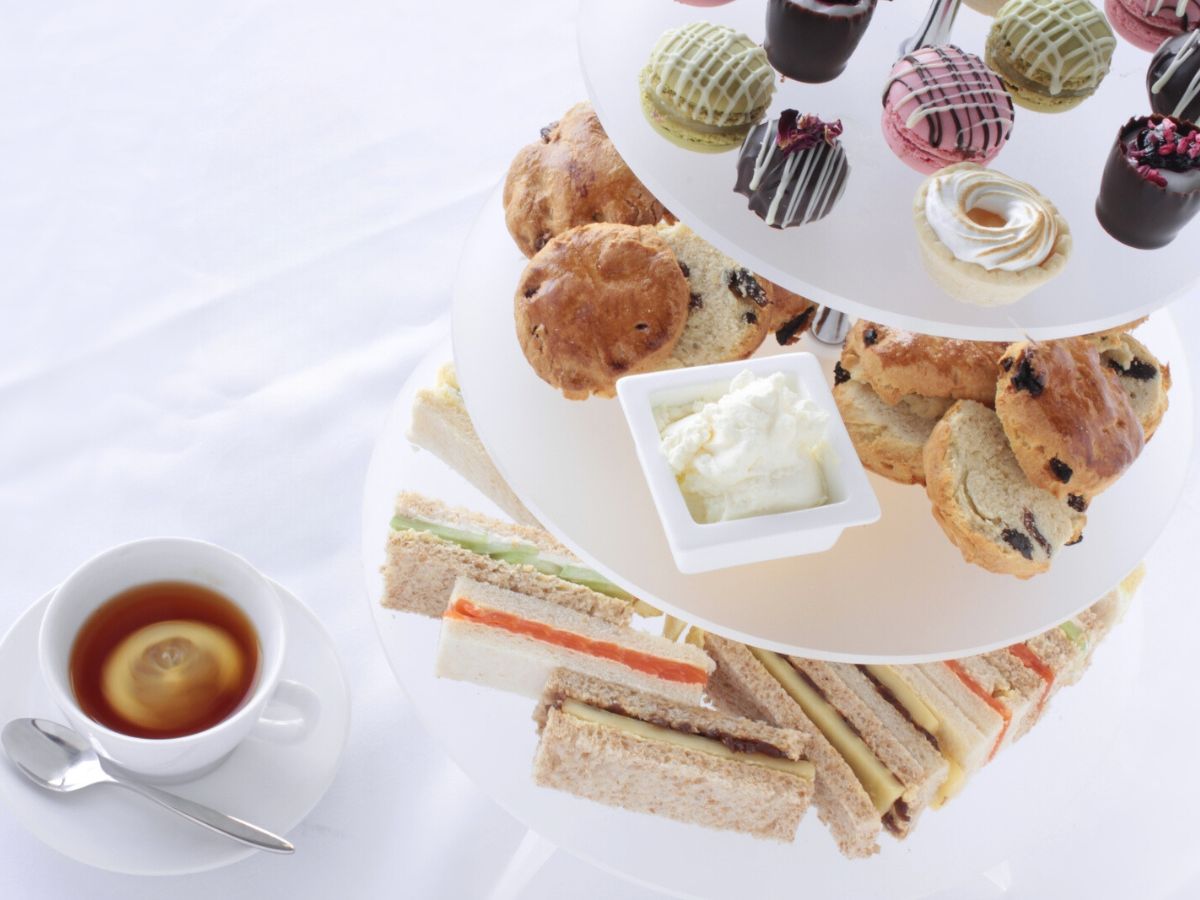 Traditional afternoon tea set with sandwiches, scones, and assorted pastries on a tiered stand, accompanied by a cup of tea.