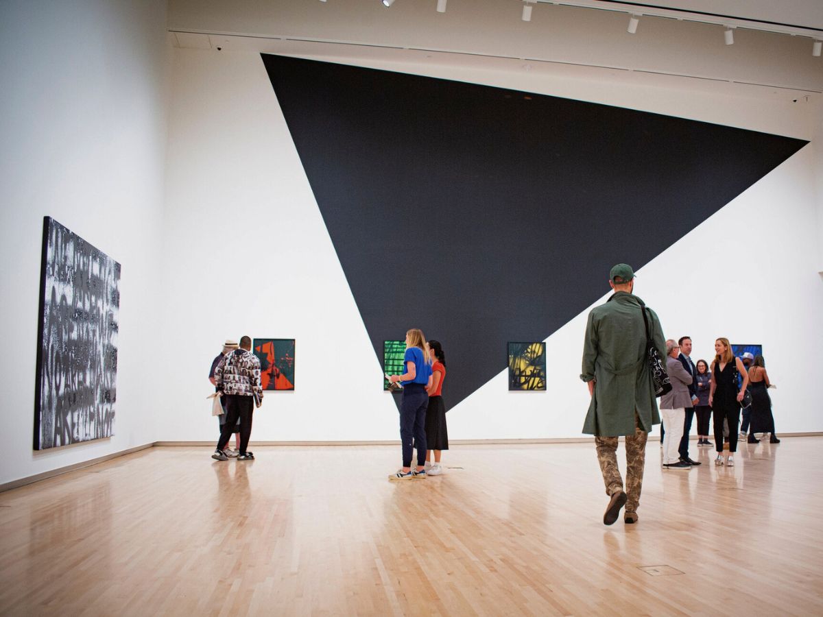 Visitors viewing modern art exhibits in a gallery with a large geometric shape on the wall.