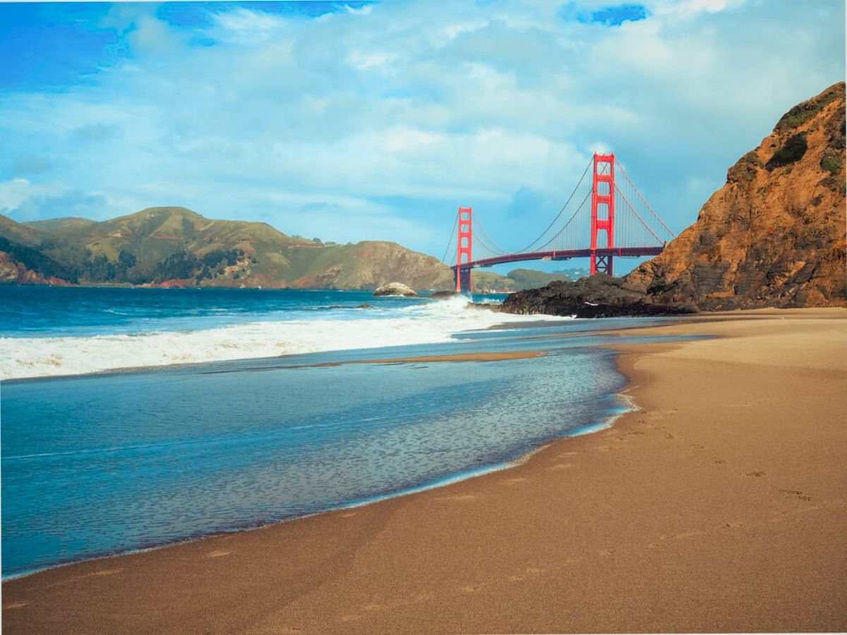 The golden gate bridge viewed from a sandy beach with gentle waves approaching the shore.