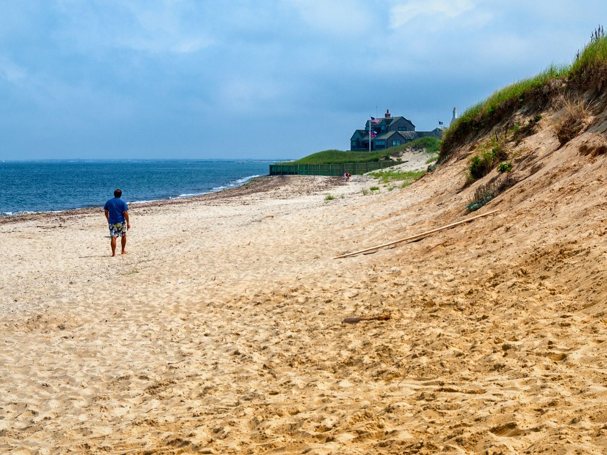 A person walking on a sandy beach in Nantucket, with a house on a grassy hill in the background under a blue sky.