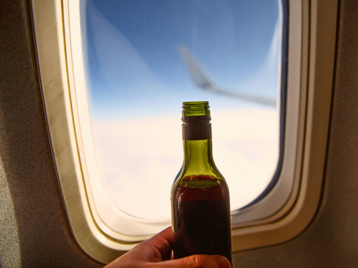A hand holding a small bottle of red wine next to an airplane window, with clouds and blue sky visible outside.