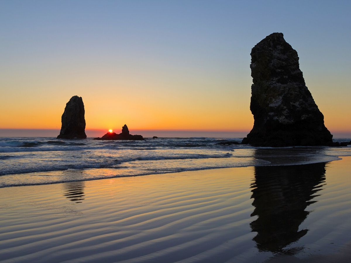 Sunset at Cannon Beach with silhouettes of large rock formations in the sea, reflecting on the wet sand.