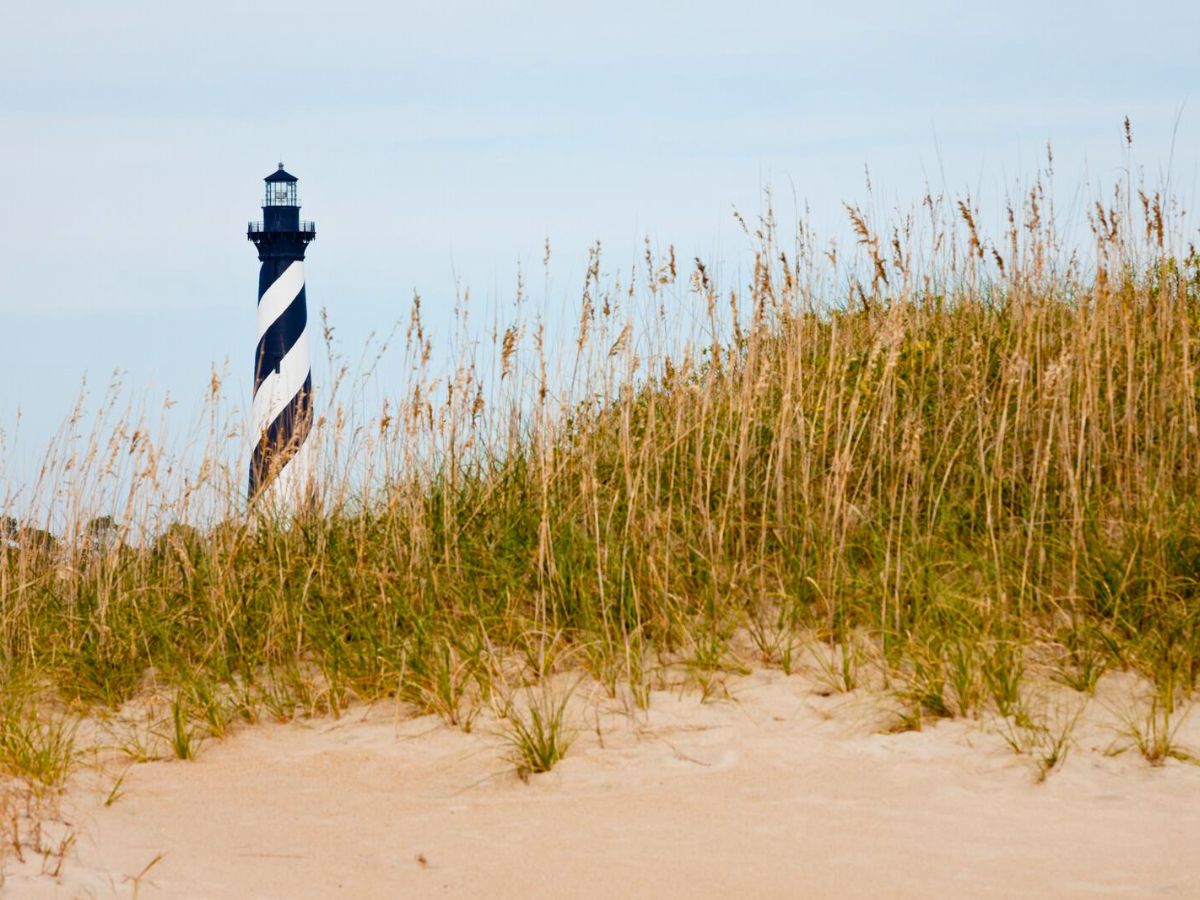 A lighthouse with black and white stripes seen beyond a sandy beach covered with tall grass.