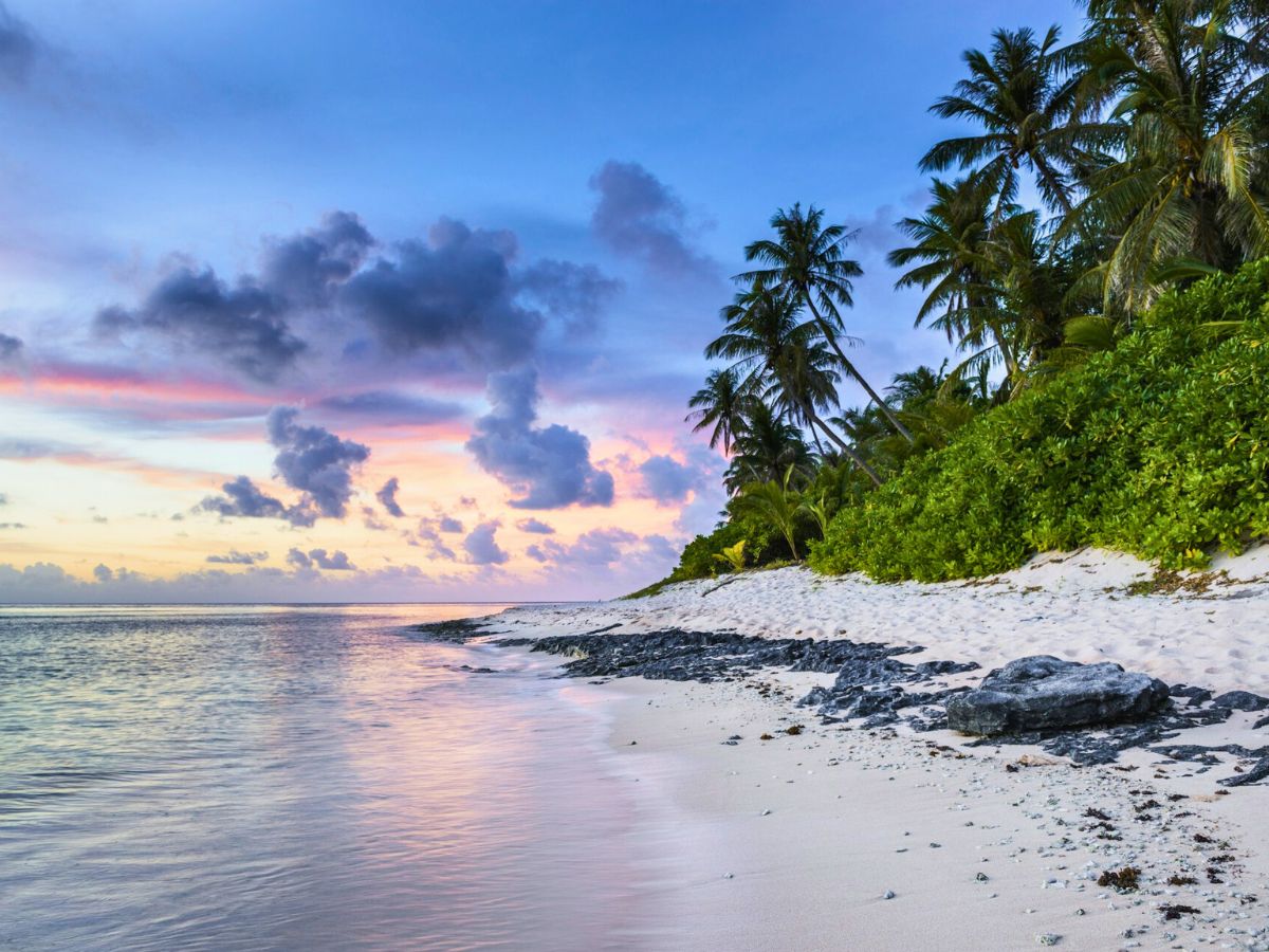 A serene tropical beach at sunset with colorful clouds, palm trees, white sands, and gentle waves.