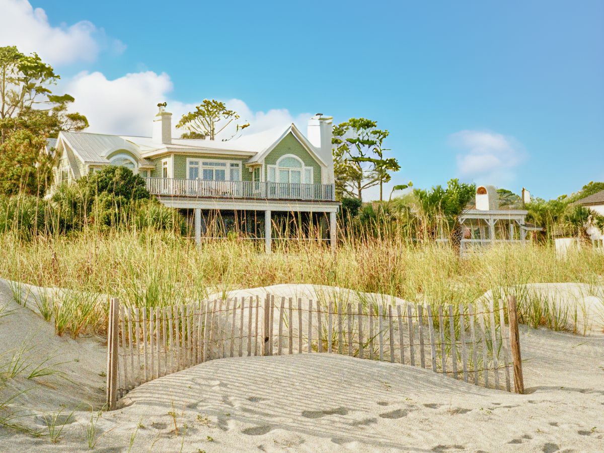 A large beachfront house with a balcony, surrounded by sand dunes and tall grass under a clear blue sky.