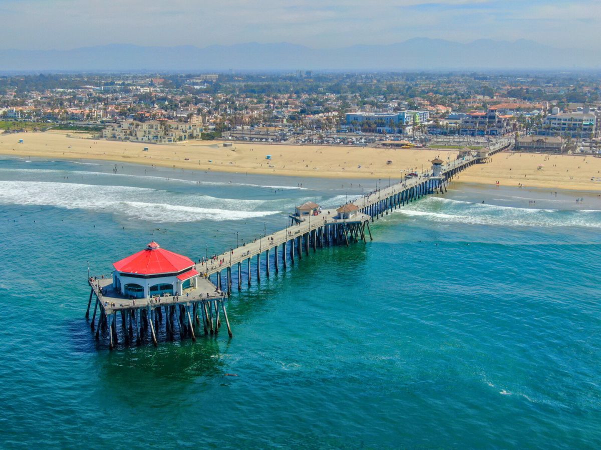 Aerial view of a pier with a red-roofed structure extending into the ocean, with a beach and coastal town in the background.
