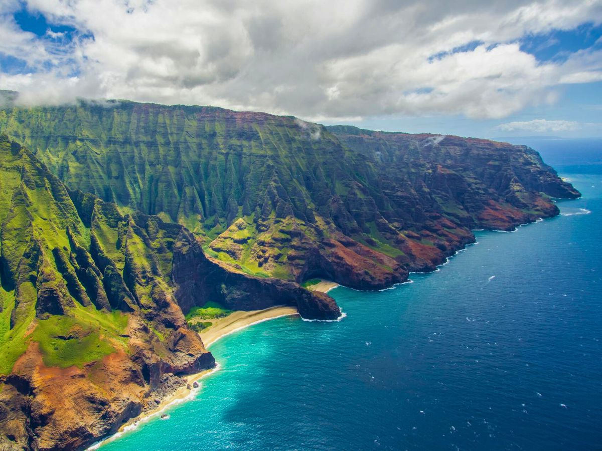 Aerial view of a rugged Kauai coastline with steep, lush cliffs and a small beach next to a turquoise ocean.
