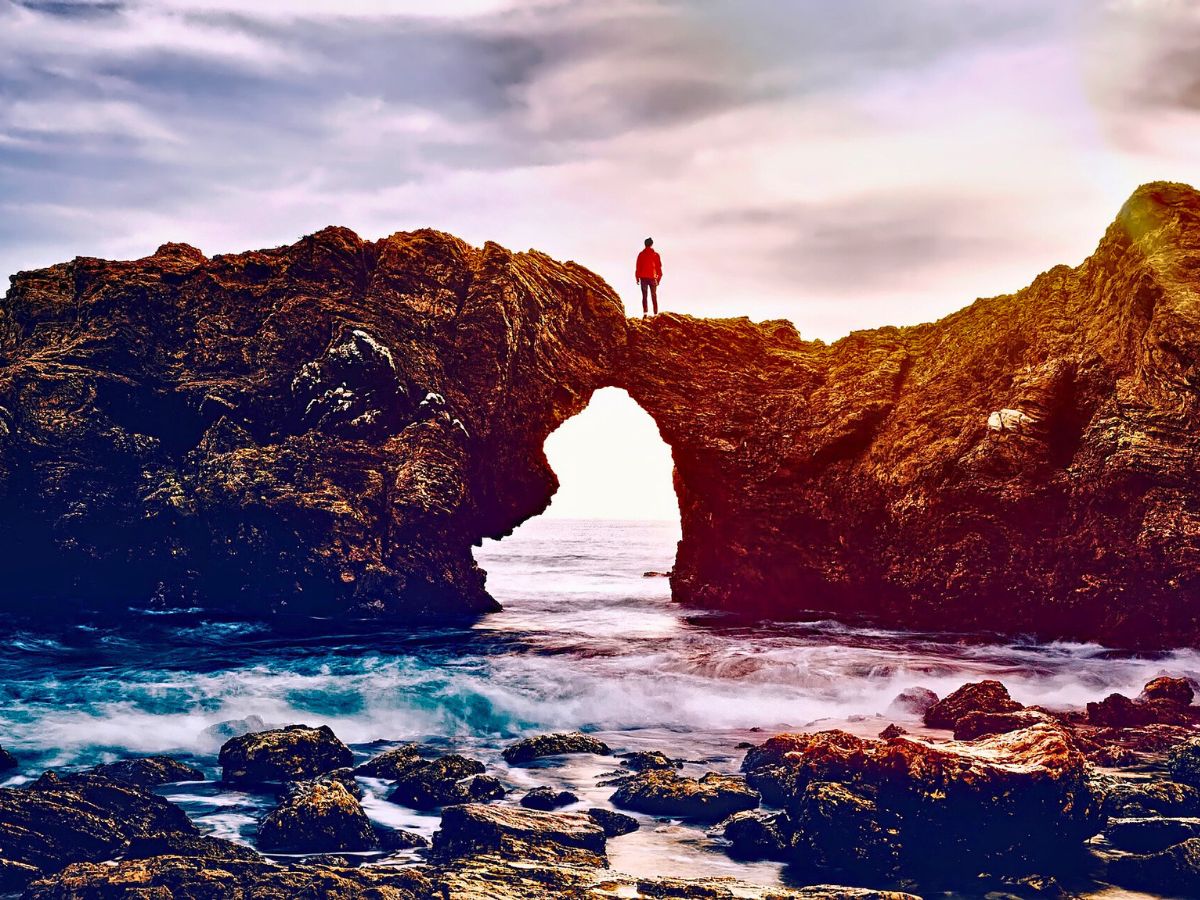 Person standing atop a sea arch off Newport Beach at sunset.