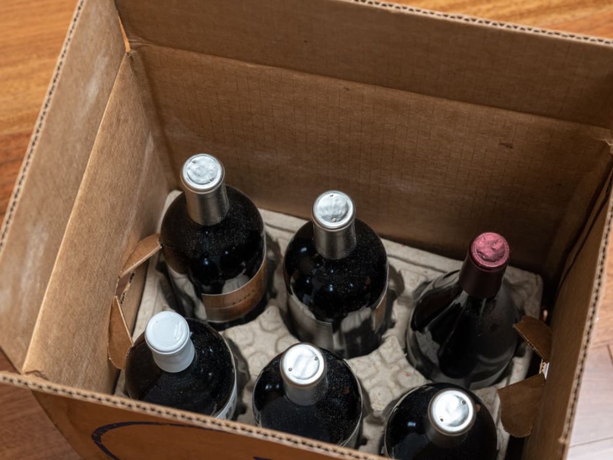 Open cardboard box with six bottles of wine, some with dark and one with a red cap, viewed from above on a wooden floor.