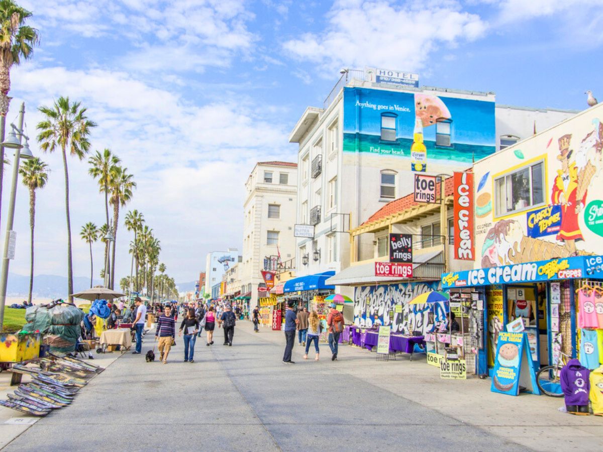 Bustling beachfront walkway with colorful storefronts and palm trees under a partly cloudy sky.