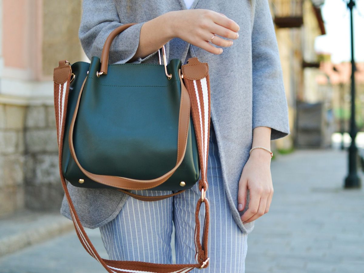 Woman holding a stylish green and brown handbag on a sunny street, wearing a grey blazer and striped trousers.