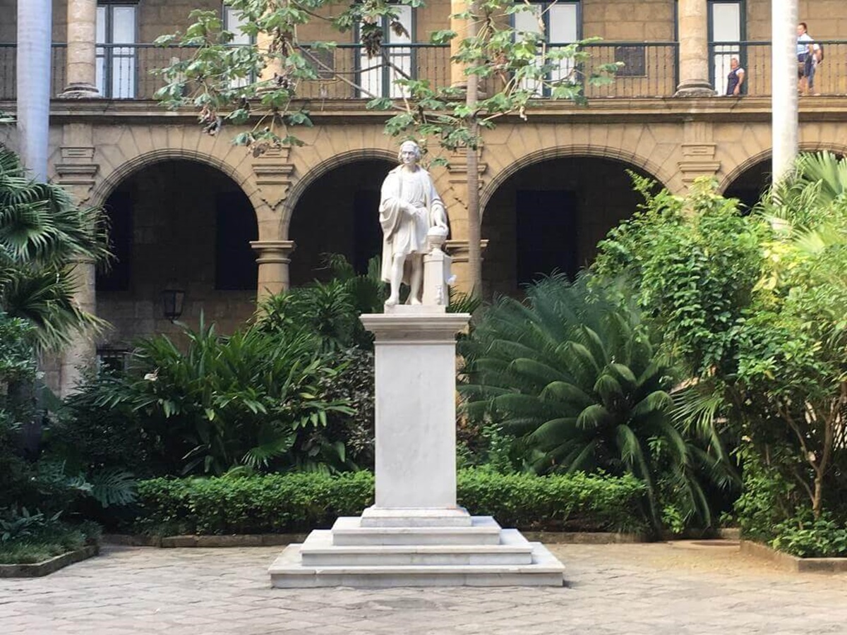 A white marble statue of a standing figure on a pedestal is situated in a lush garden courtyard with arched building in the background.
