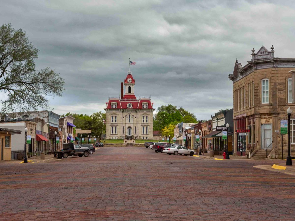A historic building with a clock tower stands at the end of a cobblestone street lined with various storefronts and parked vehicles under a cloudy sky.
