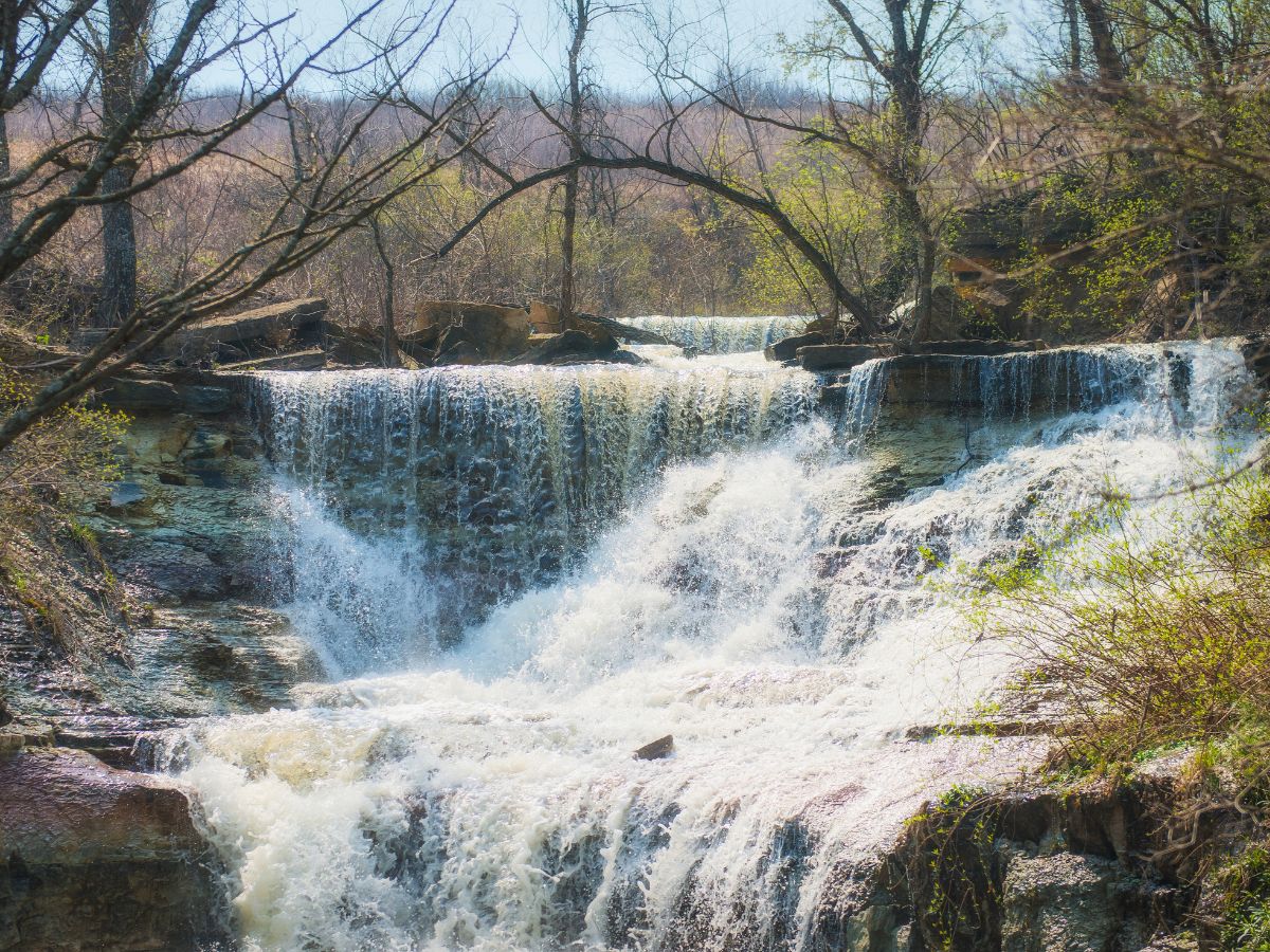 A waterfall flows over a rocky ledge surrounded by leafless trees and sparse vegetation. The water cascades down in multiple streams, creating a misty effect at the base.