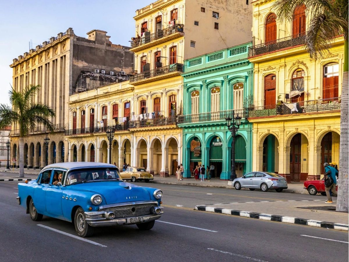 A vintage blue car drives past colorful colonial buildings along a street lined with palm trees in Havana, Cuba.