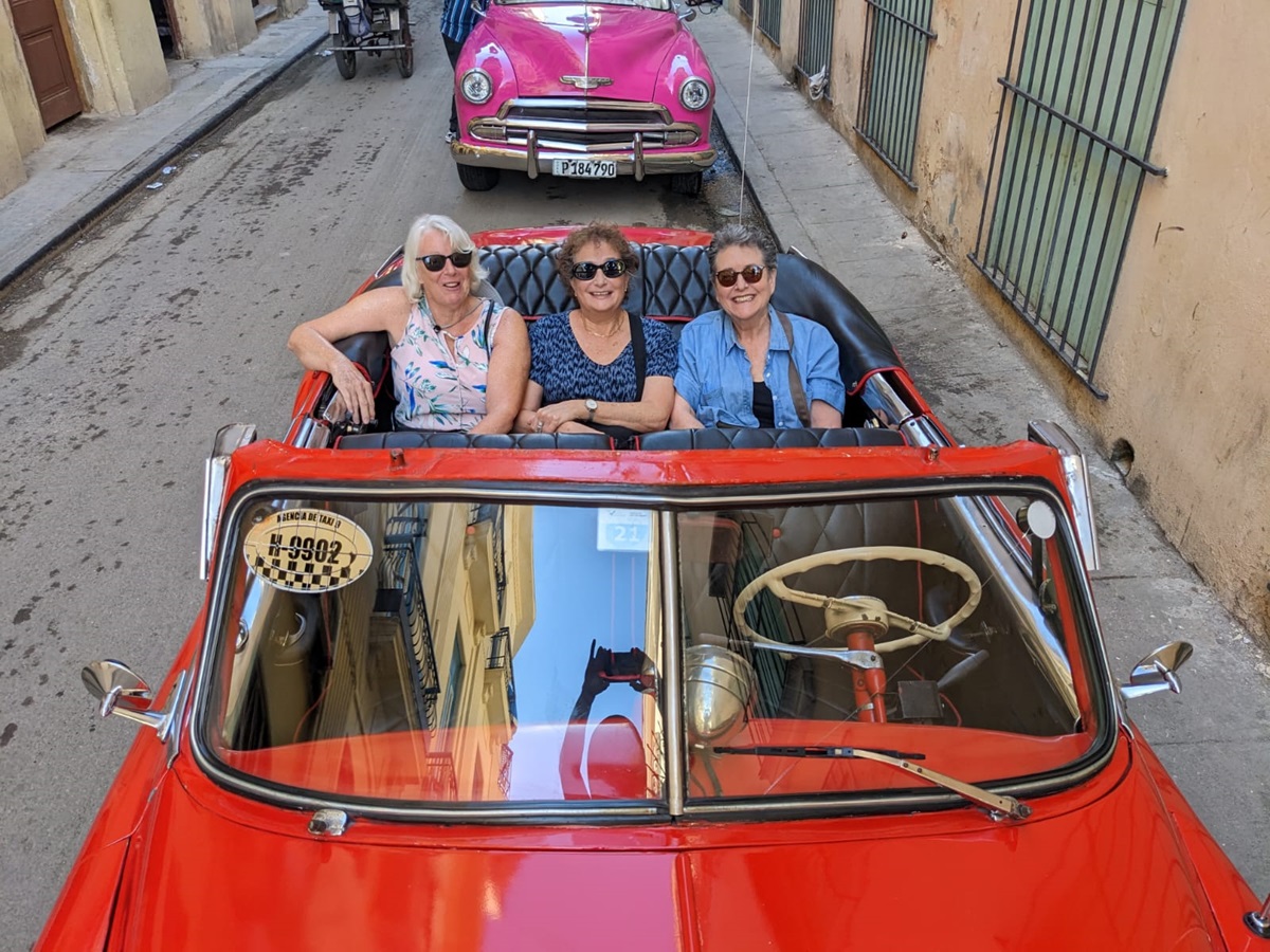Three women wearing sunglasses sit in a red convertible car driving down a narrow street with buildings on either side. A pink car is visible in the background.