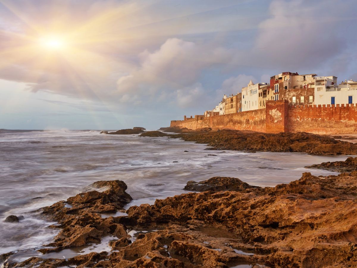 The Moroccan coastal town of Essaouira with white buildings and a brick wall overlooking a rocky shoreline.