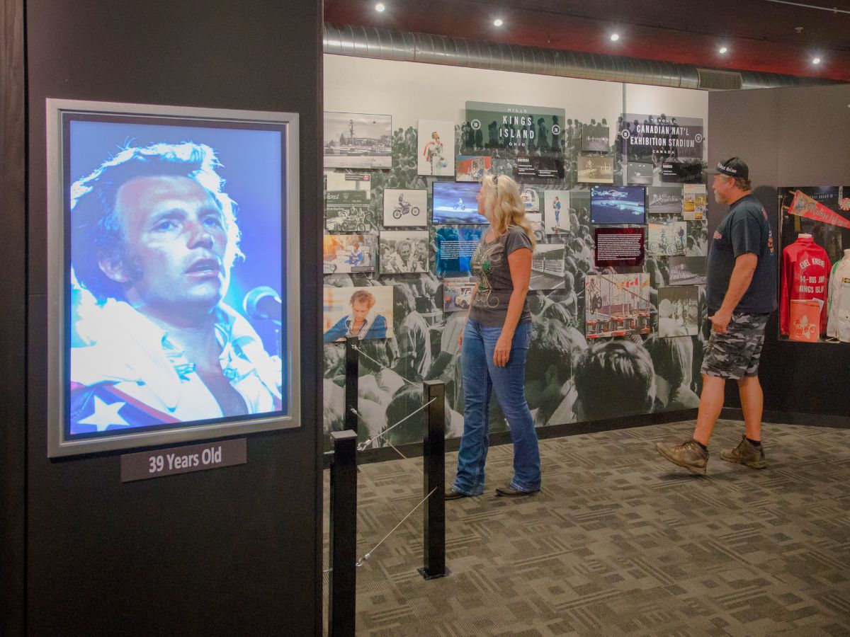 Two people observe exhibits and displays in a museum, with a large portrait of Evel Knievel on the left labeled "39 Years Old."