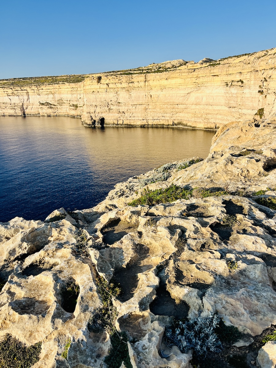 Rocky coastal cliffs with a calm sea under a clear blue sky. The limestone formations show erosion and natural patterns. Sparse vegetation is visible on the rugged terrain.