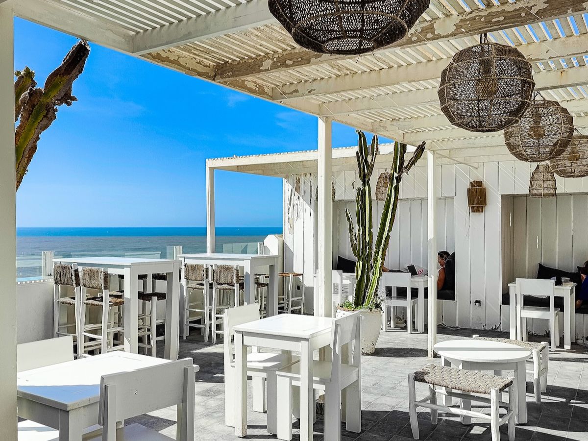 Outdoor seaside restaurant with white wooden furniture, woven hanging lamps, and cacti. The space overlooks a blue ocean under a clear sky.