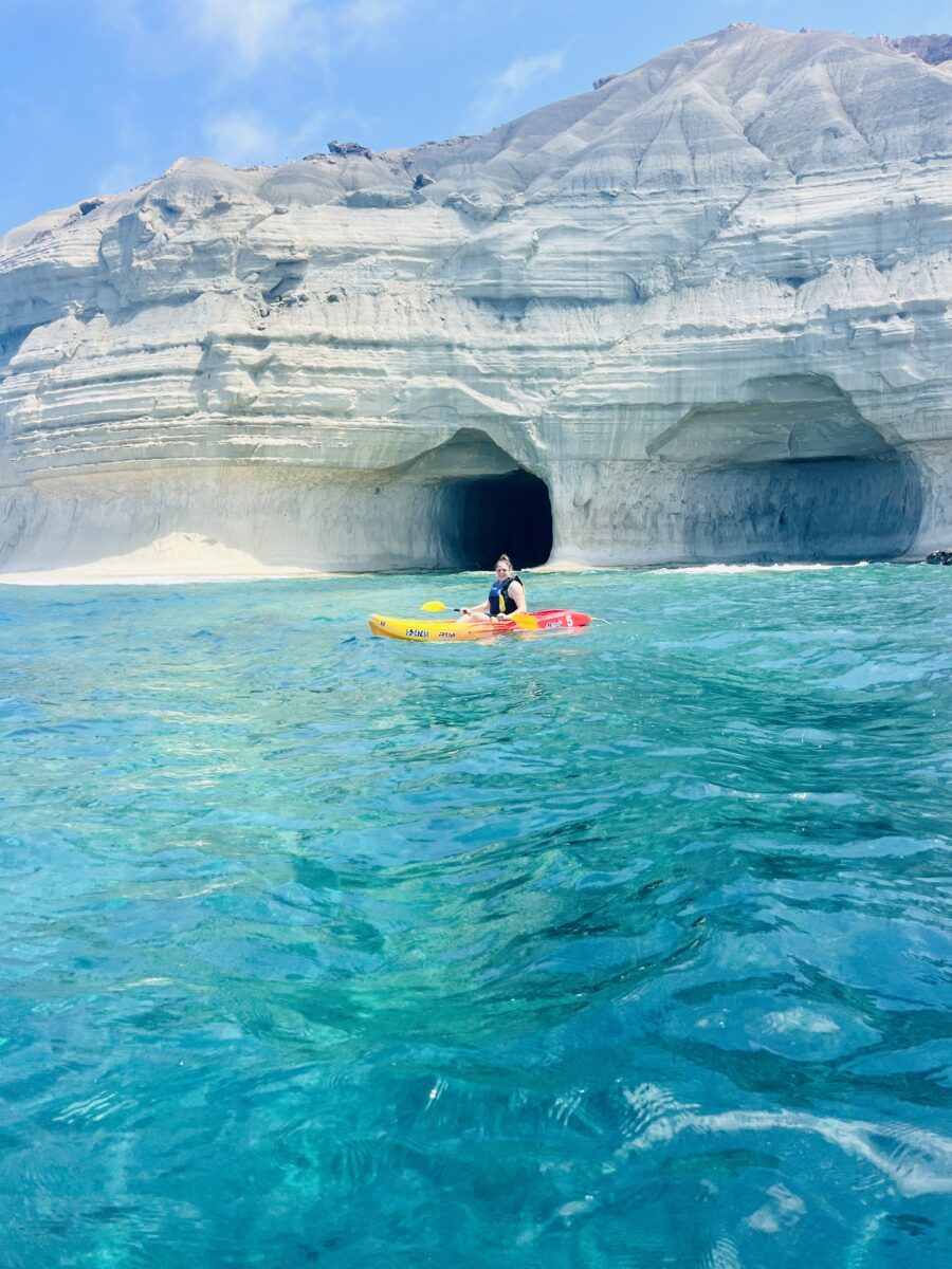 A person is kayaking in clear blue-green water near a large rocky cave formation.