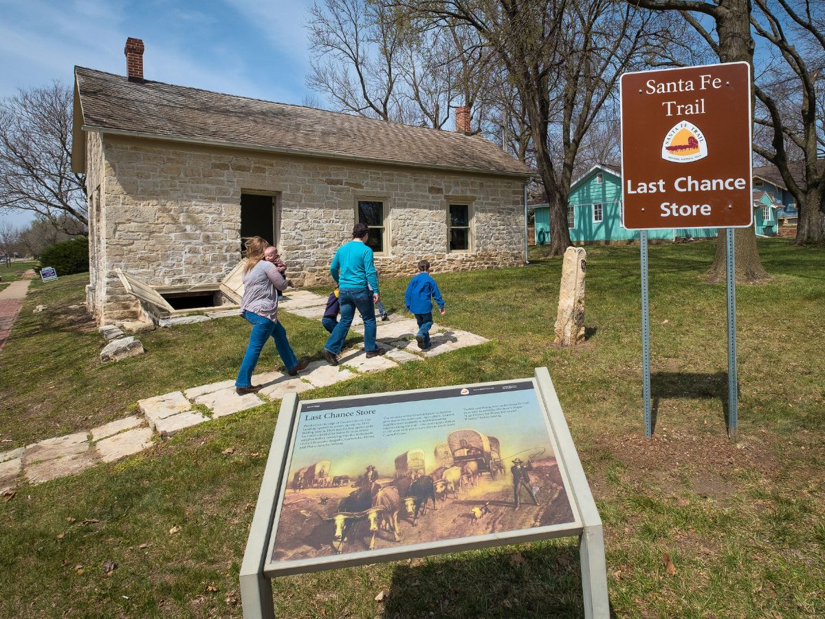 Family walking towards the Last Chance Store, a historic stone building on the Santa Fe Trail. A brown sign marks the location, and an informational plaque with an illustration stands nearby.