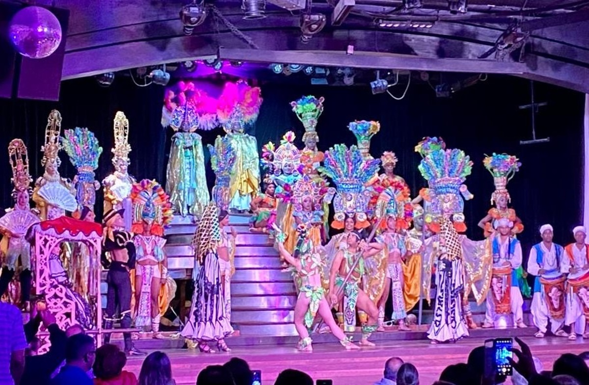 A vibrant stage performance featuring performers in elaborate, colorful costumes and headdresses. They dance on a multi-level platform, with a colorful backdrop and audience members watching.
