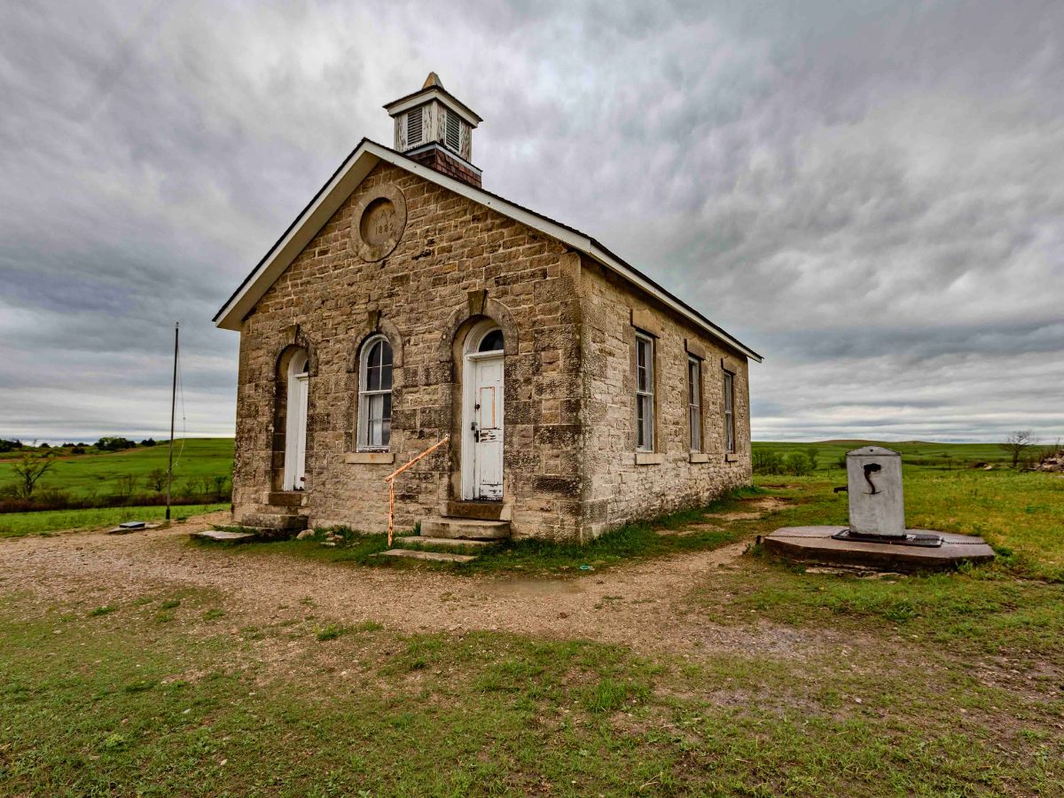 A single-story, stone building with a small cupola on the roof stands on a grassy plain under a cloudy sky. A weathered door is located at the center of the building's front.