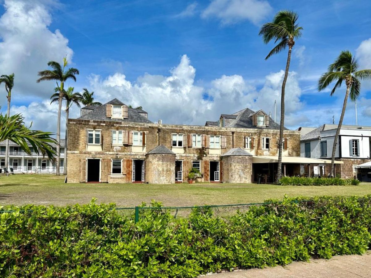 An old, large, two-story stone building with wooden shutters stands against a bright blue sky, surrounded by palm trees and manicured hedges.