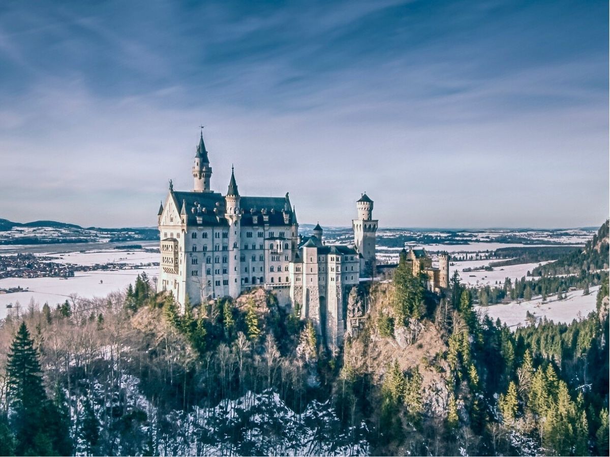 Neuschwanstein Castle in Germany stands on a hill surrounded by forest and partially snow-covered landscape under a blue sky.