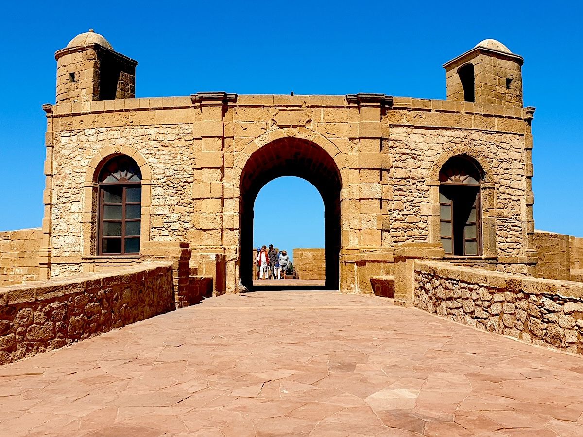 An old stone fortress with an archway and two towers under a clear blue sky, with a group of people visible through the arch.