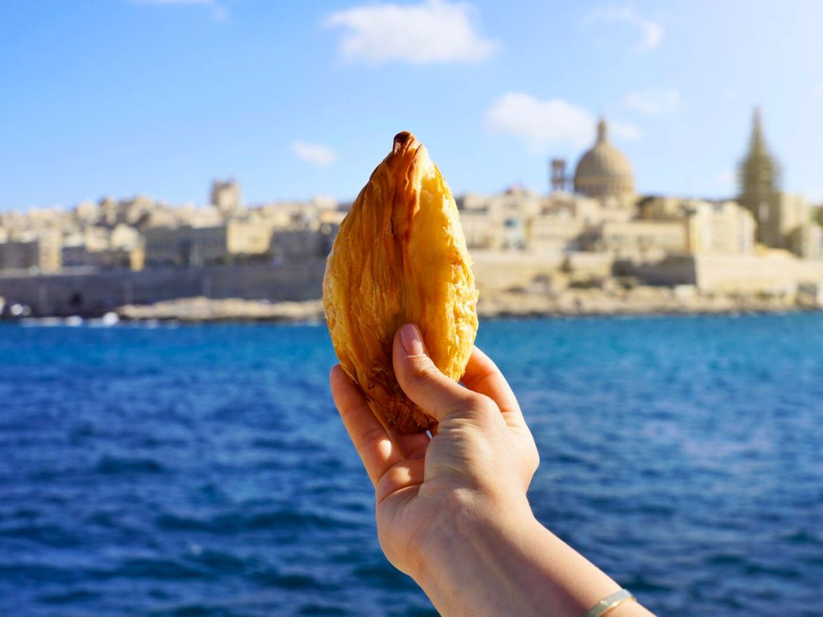 A hand holds a Maltese pastizzi pastry in front of a scenic view of a coastal city in Malta with historic buildings and a blue sky background.