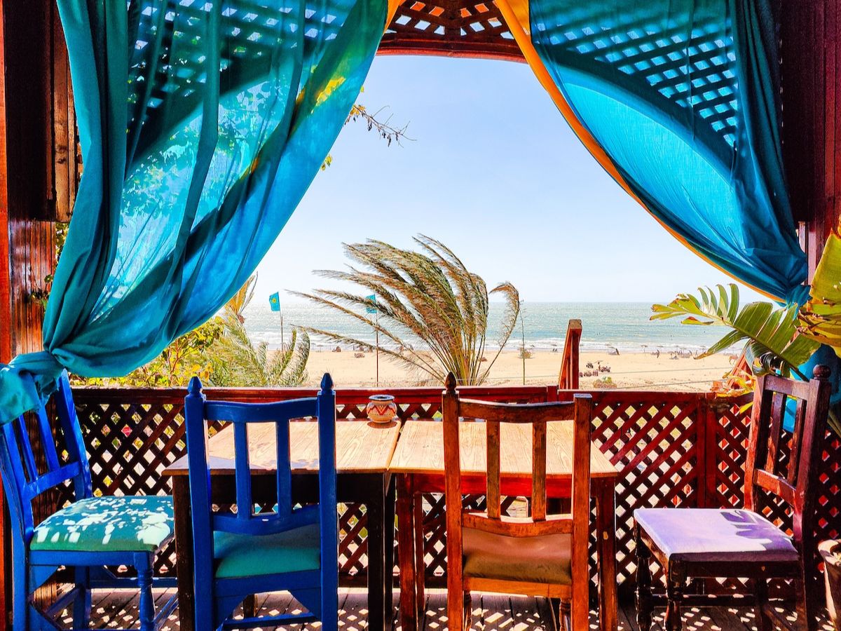 Outdoor dining area with colorful chairs and wooden table, framed by vibrant blue curtains, overlooking a beach with palm trees and clear blue sky.