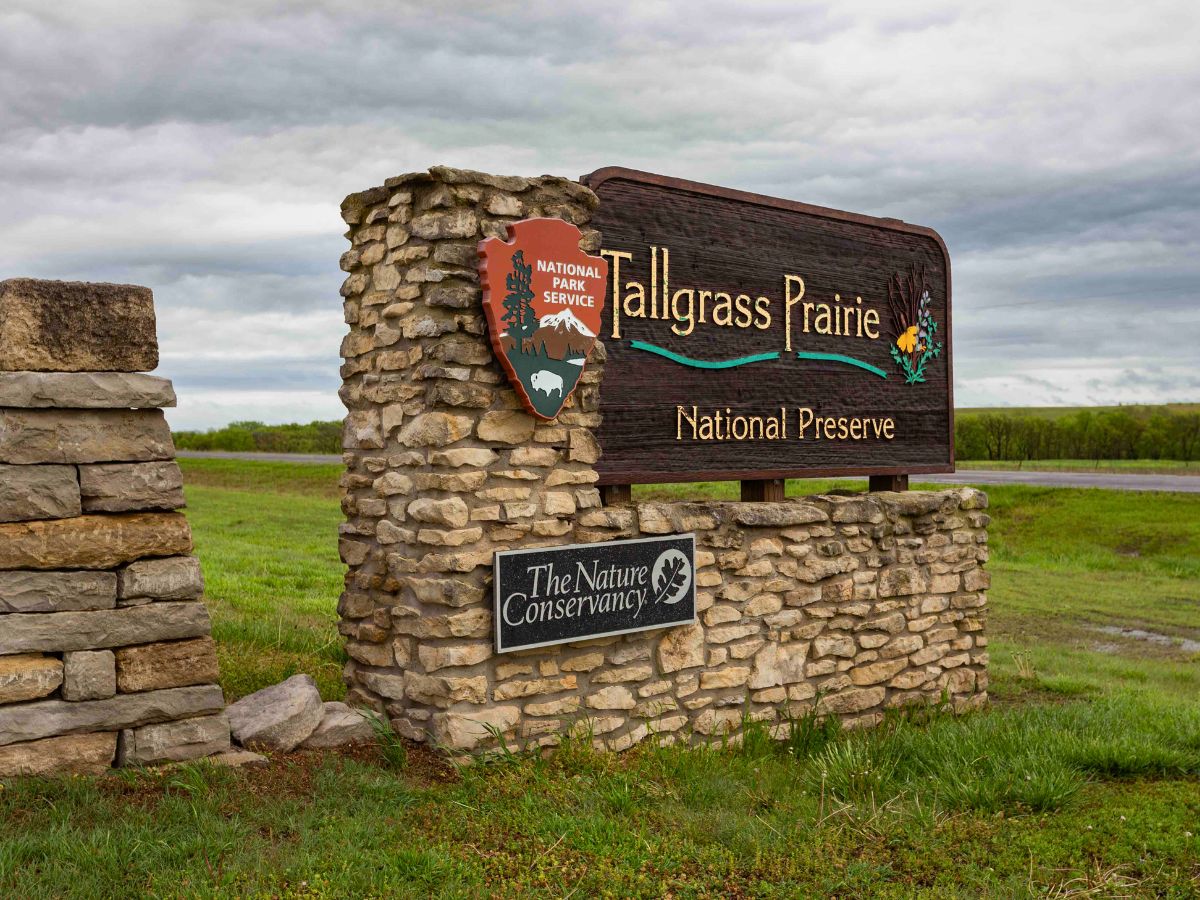 Entrance sign for Tallgrass Prairie National Preserve, featuring the National Park Service emblem, with the text "The Nature Conservancy" on a secondary plaque, set against a backdrop of grassy landscape.