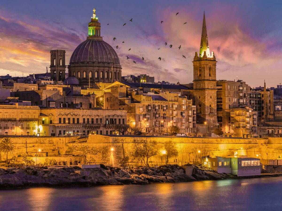 A coastal cityscape in Malta at sunset shows illuminated historic buildings, including a domed cathedral and a tall spire, with birds flying in the colorful sky and reflections in the calm water.