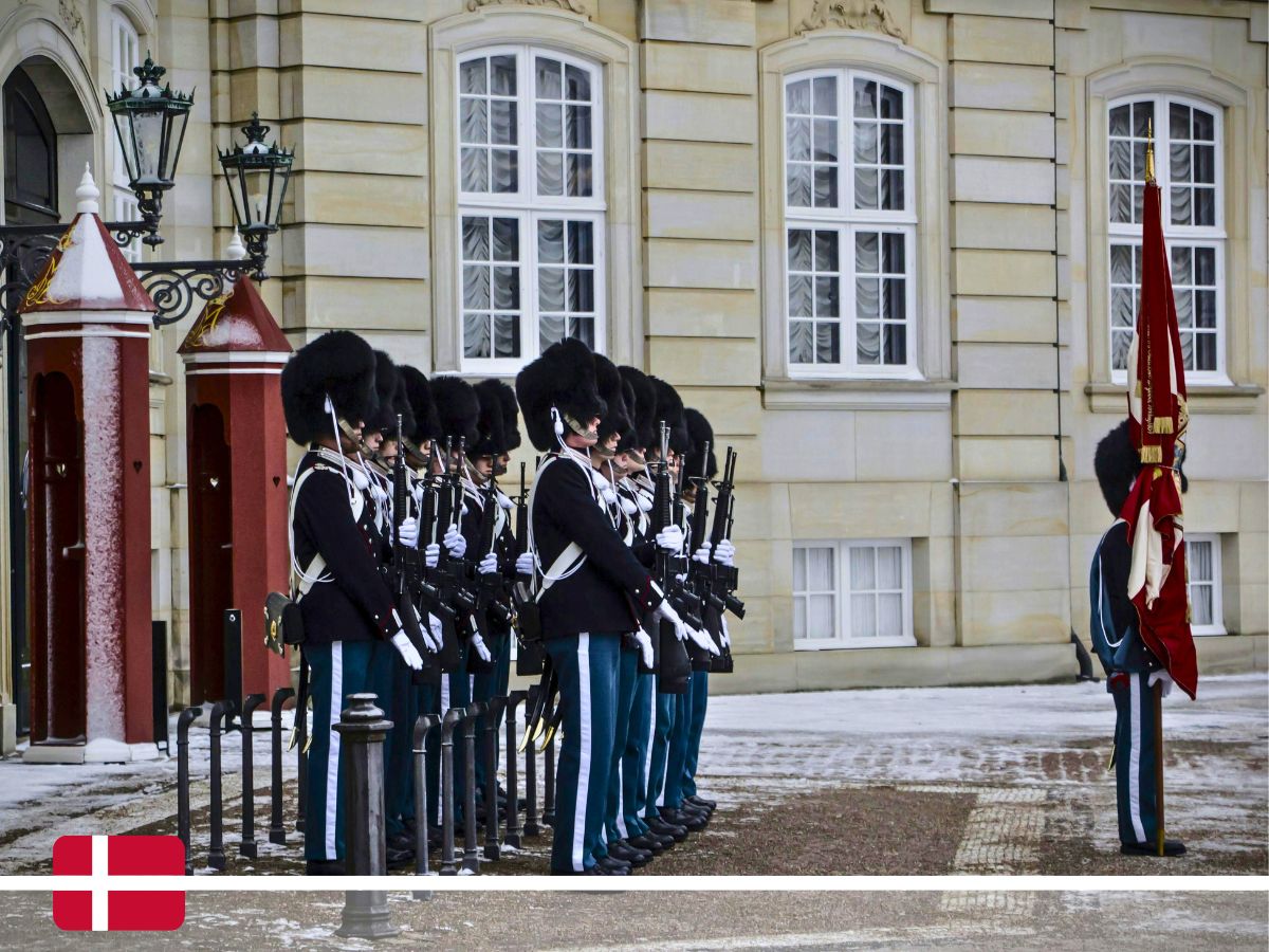 A line of guards in uniform stands at attention outside a building with large windows. One guard holds a red flag. The ground is lightly covered in snow. A Danish flag is in the lower left corner.