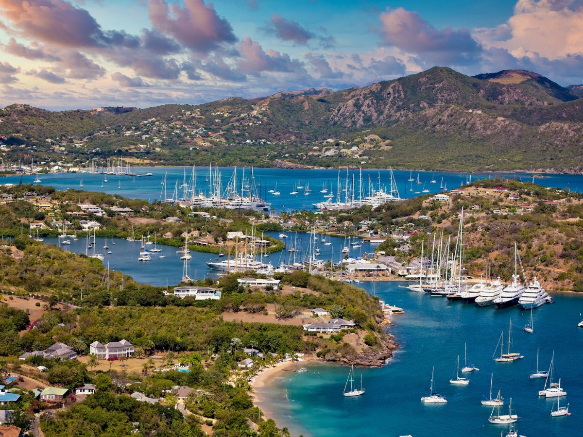 Aerial view of a coastal town with numerous sailboats and yachts anchored in a marina. The landscape is hilly, covered in greenery, and surrounded by clear blue water under a partly cloudy sky.
